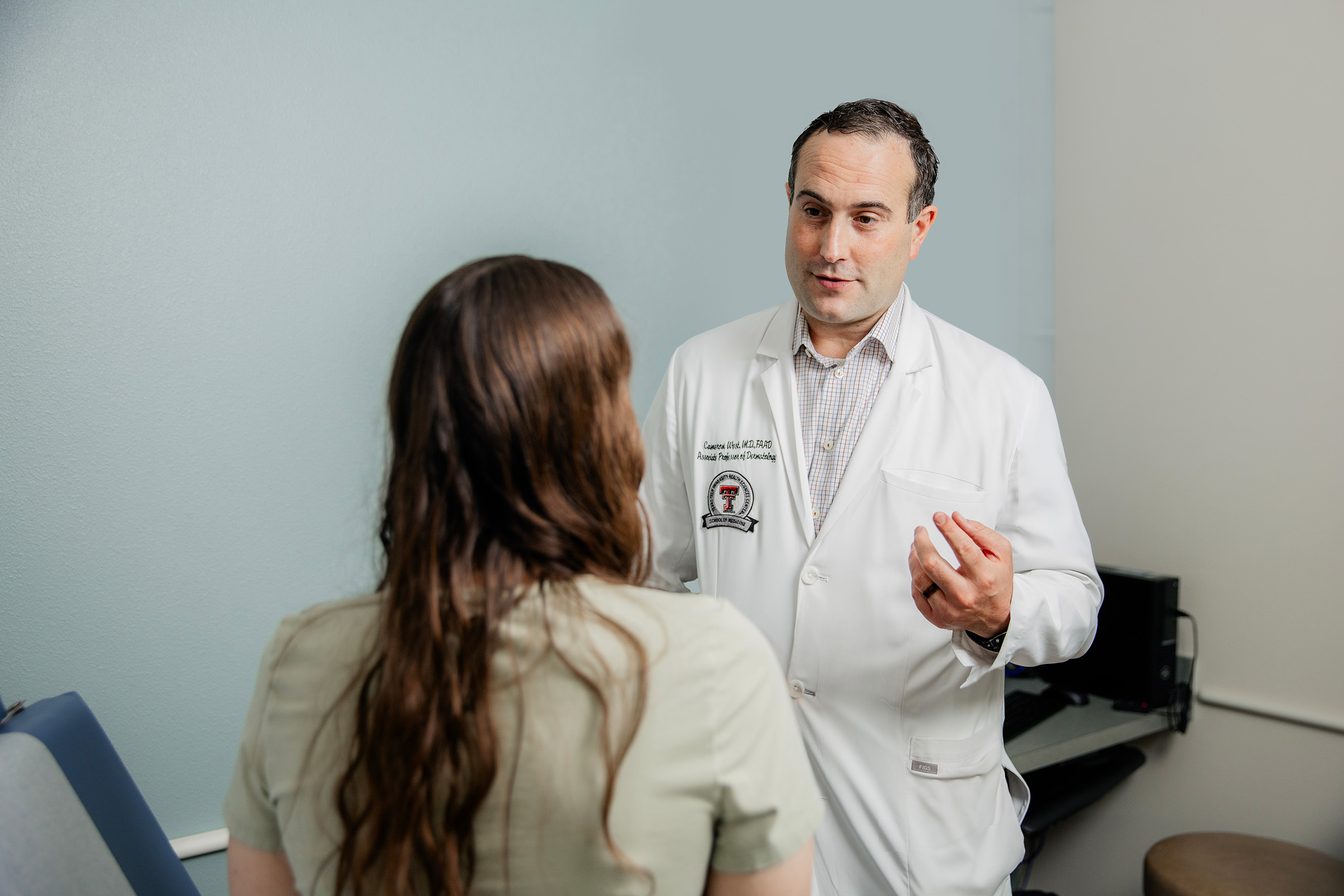 Cameron West, MD, in a clinic room speaking to patient sitting on exam table. 