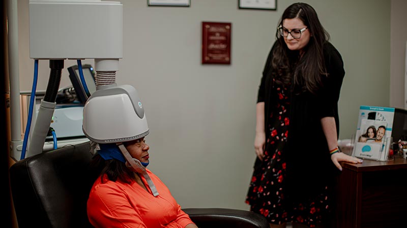 patient sitting in a chair with a tms machine on their head looking at a staff member